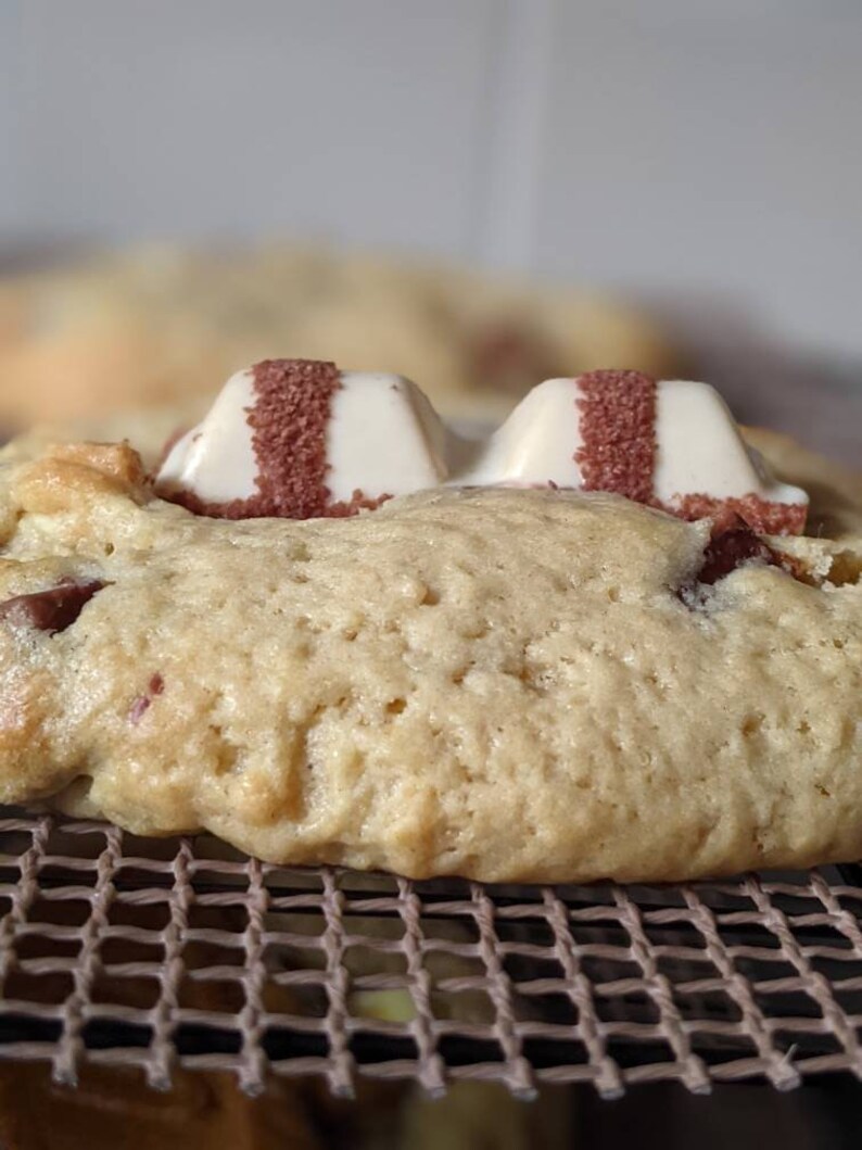 May include: Close-up of a chocolate chip cookie topped with two white chocolate squares, each with a red stripe. The cookie is resting on a brown wire rack.