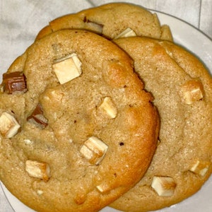 May include: Three golden brown chocolate chip cookies with white chocolate chunks on a white surface.