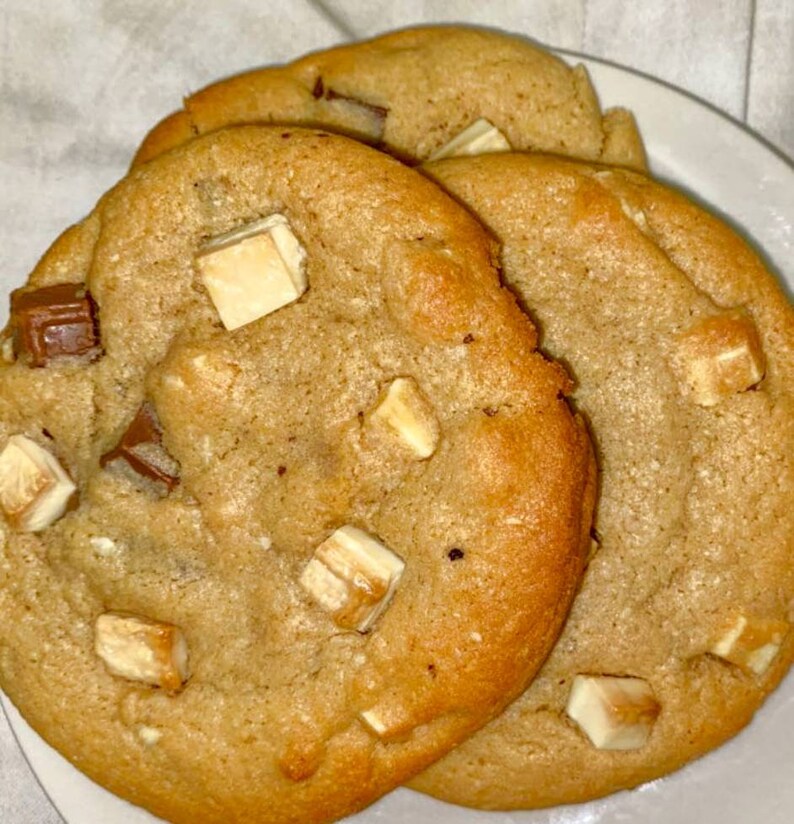 May include: Three golden brown chocolate chip cookies with white chocolate chunks on a white surface.