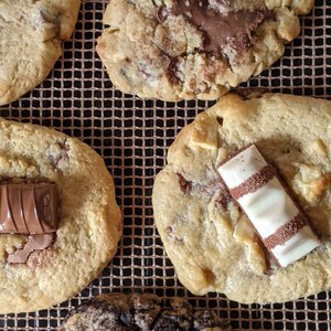 May include: Four chocolate chip cookies with different toppings. One cookie has a chocolate bar on top, another has a white chocolate bar, another has chocolate chips, and the last one has a chocolate bar and chocolate chips.