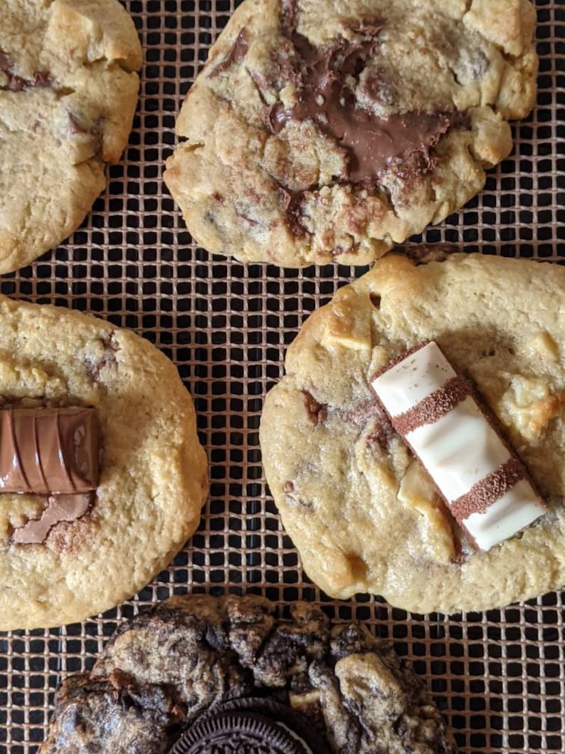 May include: Four chocolate chip cookies with different toppings. One cookie has a chocolate bar on top, another has a white chocolate bar, another has chocolate chips, and the last one has a chocolate bar and chocolate chips.