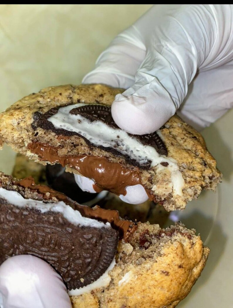 May include: A chocolate chip cookie with a chocolate and white frosting filling and an Oreo cookie inside. The cookie is broken in half, revealing the filling and Oreo cookie.