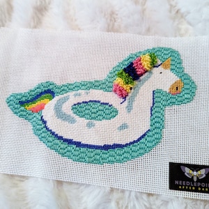 May include: A white needlepoint canvas with a colorful unicorn pool float design. The float is turquoise blue with white accents and a rainbow tail. The unicorn has a pink, yellow, green, and blue mane.