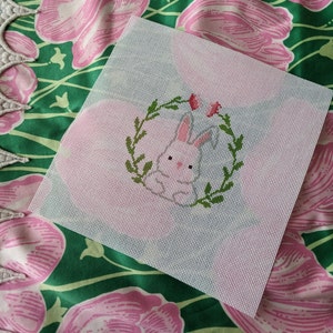 May include: White canvas with a pink bunny rabbit in a green wreath with pink flowers. The bunny is in the center of the canvas and is surrounded by the wreath. The wreath is made of green leaves and has pink flowers on it.