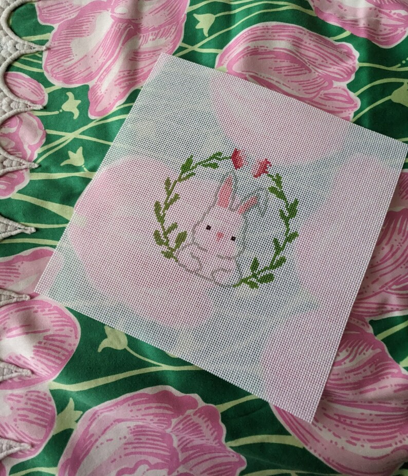 May include: White canvas with a pink bunny rabbit in a green wreath with pink flowers. The bunny is in the center of the canvas and is surrounded by the wreath. The wreath is made of green leaves and has pink flowers on it.