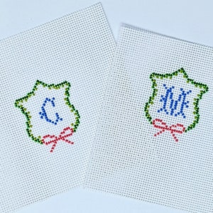 May include: Two white canvas needlepoint ornaments with green, blue, and red embroidery. The ornaments feature a star-shaped frame with a monogram in the center and a red bow.