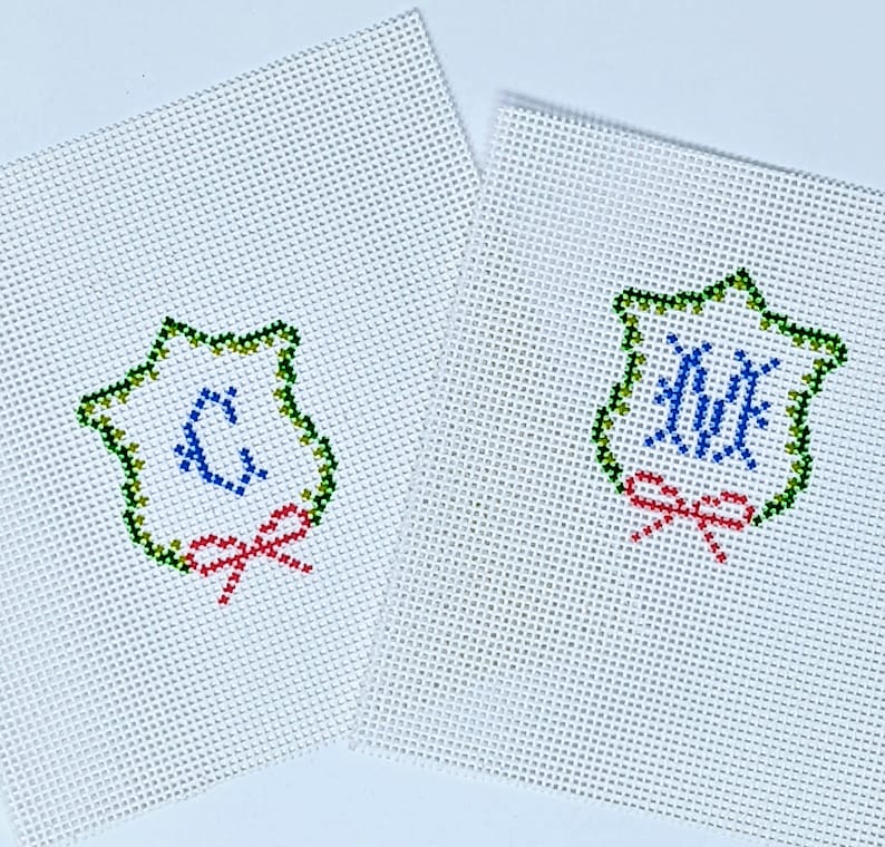 May include: Two white canvas needlepoint ornaments with green, blue, and red embroidery. The ornaments feature a star-shaped frame with a monogram in the center and a red bow.