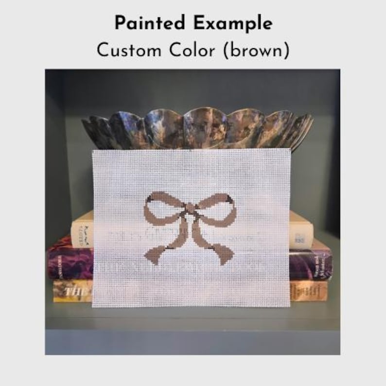 Puede incluir: Un lienzo de punto de aguja con un dise&ntilde;o de lazo marr&oacute;n. El lienzo se exhibe en una estanter&iacute;a con libros y un cuenco decorativo. El texto "Painted Example Custom Color (brown)" est&aacute; en la parte superior.