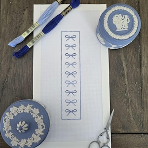 May include: A needlepoint project with a white canvas and a blue bow design. Two spools of embroidery thread in light and dark blue, two blue decorative boxes, and a pair of scissors are also present. The items are arranged on a wooden surface.