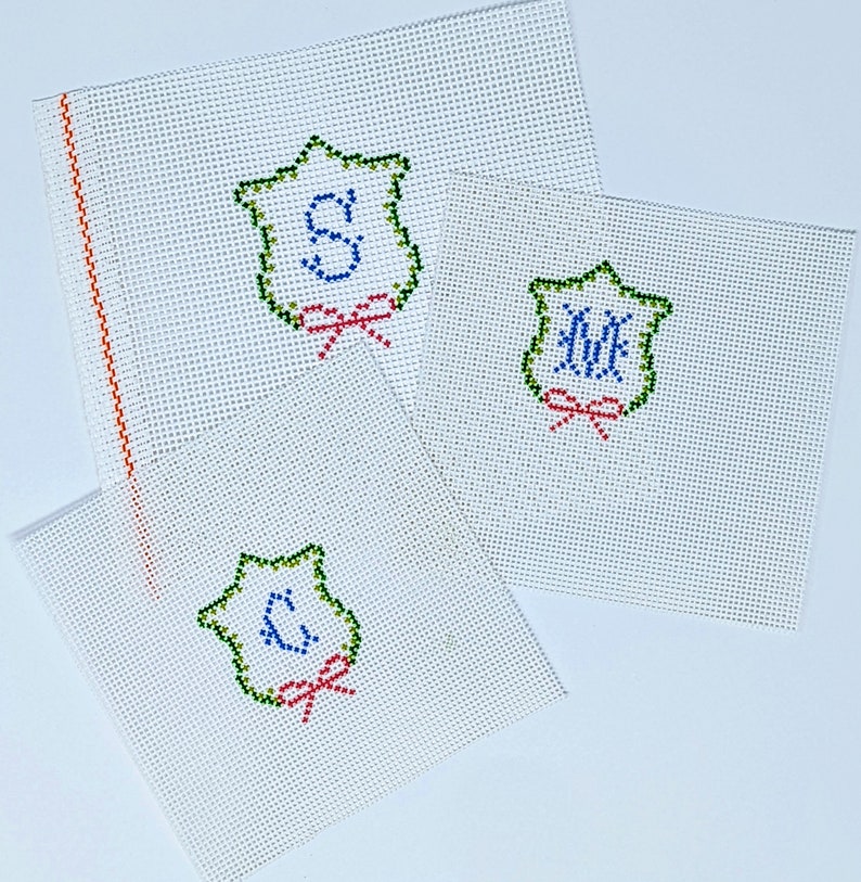 May include: Three white canvas panels with green, blue, and red embroidered designs. Each panel features a shield-shaped design with a letter inside. The letters are "S", "M", and "C".