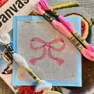 Puede incluir: Un lienzo de punto de aguja con un dise&ntilde;o de lazo rosa, rodeado por un borde azul claro. Tres madejas de hilo de bordar en tonos rosa y blanco est&aacute;n dispuestas alrededor del lienzo, listas para coser. La palabra "Canvas" es parcialmente visible en el fondo.