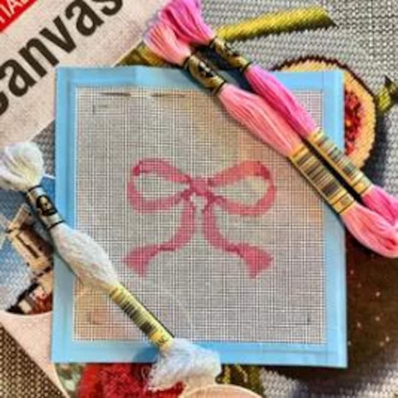 Puede incluir: Un lienzo de punto de aguja con un dise&ntilde;o de lazo rosa, rodeado por un borde azul claro. Tres madejas de hilo de bordar en tonos rosa y blanco est&aacute;n dispuestas alrededor del lienzo, listas para coser. La palabra "Canvas" es parcialmente visible en el fondo.