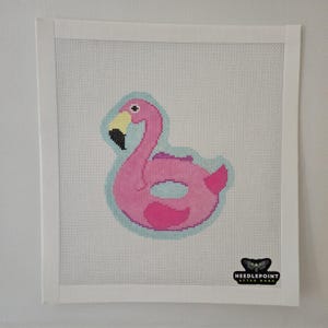 Puede incluir: Lienzo de punto de aguja con un diseño de flotador de flamenco rosa. El flamenco tiene un pico amarillo, detalles en negro y está delineado en azul claro. El lienzo es cuadrado y tiene un borde blanco. El logotipo de Needlepoint After Dark está en la esquina inferior derecha.