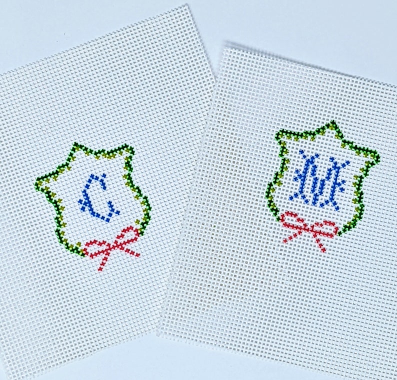 May include: Two white canvas needlepoint ornaments with green, blue, and red embroidery. The ornaments feature a star-shaped frame with a monogram in the center and a red bow.
