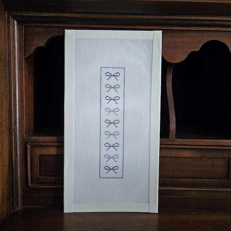 May include: A rectangular needlepoint piece with a white border. The needlepoint features a series of blue bow designs in a vertical pattern. The background is a light blue grid.