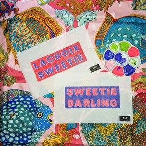 May include: Two white canvas cross stitch patterns with colourful embroidery floss. The top pattern reads "LACROIX SWEETIE" in pink and purple lettering. The bottom pattern reads "SWEETIE DARLING" in purple and pink lettering. The patterns are on a pink and blue patterned fabric with fish and coral designs.