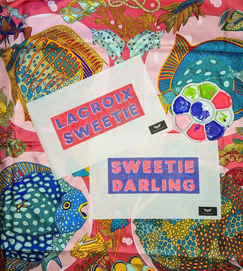 May include: Two white canvas cross stitch patterns with colourful embroidery floss. The top pattern reads "LACROIX SWEETIE" in pink and purple lettering. The bottom pattern reads "SWEETIE DARLING" in purple and pink lettering. The patterns are on a pink and blue patterned fabric with fish and coral designs.