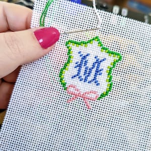 May include: A white Aida cloth with a green, blue, and pink embroidered design of a shield with the letter "M" inside and a pink bow at the bottom.