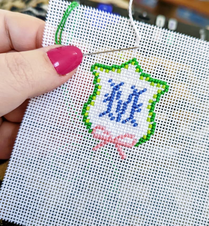 May include: A white Aida cloth with a green, blue, and pink embroidered design of a shield with the letter "M" inside and a pink bow at the bottom.