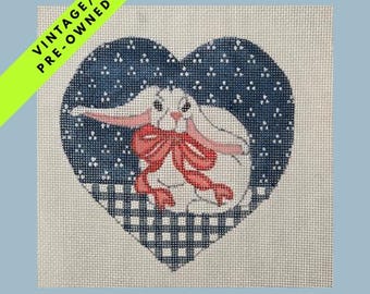 May include: Vintage pre-owned needlepoint canvas featuring a white bunny with pink ears and a red bow, set against a blue heart-shaped background with a dotted pattern and a gingham border. The canvas has a light beige color.