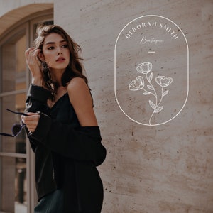 Puede incluir: Una mujer con un top y una chaqueta negros se para frente a una pared con un logotipo ovalado blanco que dice "Deborah Smith Boutique" con un dise&ntilde;o floral estilizado.