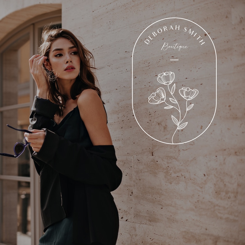 Puede incluir: Una mujer con un top y una chaqueta negros se para frente a una pared con un logotipo ovalado blanco que dice "Deborah Smith Boutique" con un dise&ntilde;o floral estilizado.