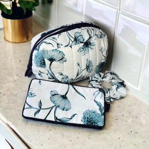May include: A set of three white quilted bags with a blue floral pattern. The bags are a large makeup bag, a smaller pouch, and a hair scrunchie.