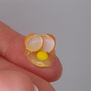May include: A miniature fried egg, with a bright yellow yolk and a translucent white, with two halves of the eggshell. The egg sits on a clear, amber-coloured base, creating a whimsical, food-themed miniature.