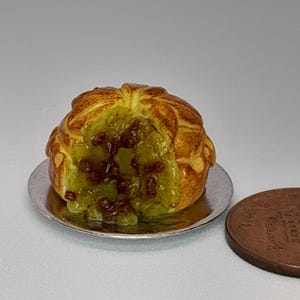 May include: A miniature baked pastry with a golden-brown, scored crust and a visible green and brown fruit filling, resting on a small silver-coloured circular plate next to a copper-coloured coin for scale, showing its small size.