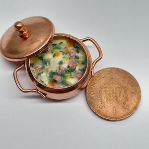 May include: A miniature copper pot with a lid, filled with a creamy soup with green herbs and small pieces of meat. A copper-coloured British one pence coin is next to the pot.
