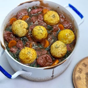 May include: A small white pot with blue handles, filled with a stew. The stew includes meat pieces, orange carrot slices, green vegetables, and six yellow dumplings. A copper coin is in the foreground, next to the pot.
