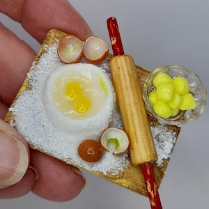 May include: A miniature kitchen scene featuring a wooden rolling pin, a small bowl of yellow sweets, and a dish with eggs. The eggs are cracked open, with yolks visible. The scene is set on a small, square wooden board.