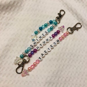 May include: Three beaded keychains with star charms. The keychains spell out the names "SHINee", "HYUNA", and "TAEYEON" in white letters on white beads. The beads are in shades of blue, purple, and pink.