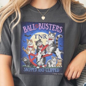 May include: Dark gray t-shirt featuring a graphic with the text "BALL BUSTERS" and "SNIPPED AND CLIPPED". The graphic depicts several cats, ghosts, and bats against a blue night sky background. The shirt is a casual, unisex style.