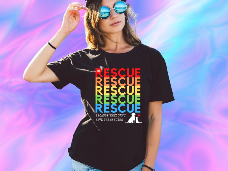 May include: A black t-shirt with a rainbow design that says "RESCUE" five times. The text is in a retro font. Below the text, it says "BECAUSE THEY CAN'T SAVE THEMSELVES" with a small white silhouette of a dog and cat.