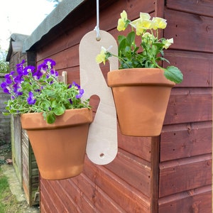 May include: Two terracotta pots with flowering plants hanging from a wooden planter. The planter is white and has a simple design. The plants are in bloom with purple and yellow flowers.