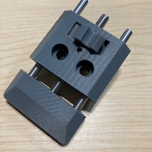 May include: A grey 3D printed clamp with two adjustable screws and a lever. The clamp is designed to hold objects securely in place.