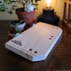 Op de afbeelding: Een licht houten snijplank met afgeronde randen en gaten die in het oppervlak zijn geboord. De plank heeft de tekst "The Wooden Rat" en een klein beeld van drie bomen gegraveerd op het oppervlak.