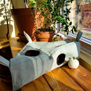 May include: A light green canvas cat tunnel with two openings and a small, white ceramic cat toy with a green plant on top. The tunnel is sitting on a wooden surface.