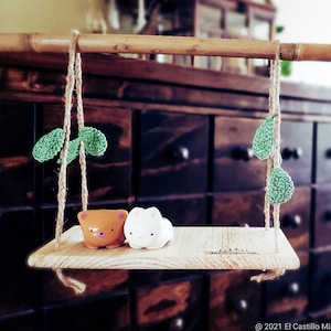 May include: A wooden swing set with two small ceramic figurines, one brown and one white, sitting on the swing. The swing is hanging from a bamboo rod and is decorated with green crocheted leaves.
