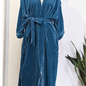 May include: A teal blue velvet robe with a long, flowing silhouette. The robe has a V-neckline, long sleeves, and a tie belt. It is made of a soft, luxurious fabric.
