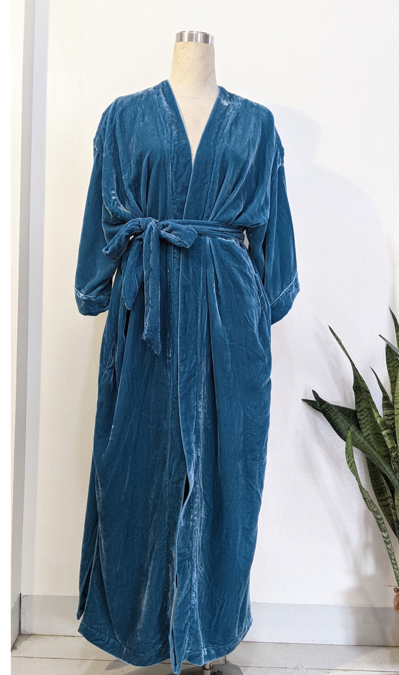 May include: A teal blue velvet robe with a long, flowing silhouette. The robe has a V-neckline, long sleeves, and a tie belt. It is made of a soft, luxurious fabric.