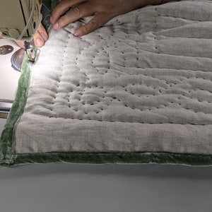 May include: A person is sewing a green velvet trim onto a light grey quilted fabric with a sewing machine. The fabric has a straight stitch pattern.
