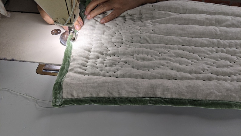 May include: A person is sewing a green velvet trim onto a light grey quilted fabric with a sewing machine. The fabric has a straight stitch pattern.