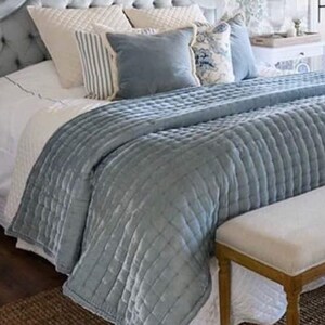 May include: A light blue quilted bedspread with a white bed and a white bench at the foot of the bed. The bed is made with white and blue patterned pillows.