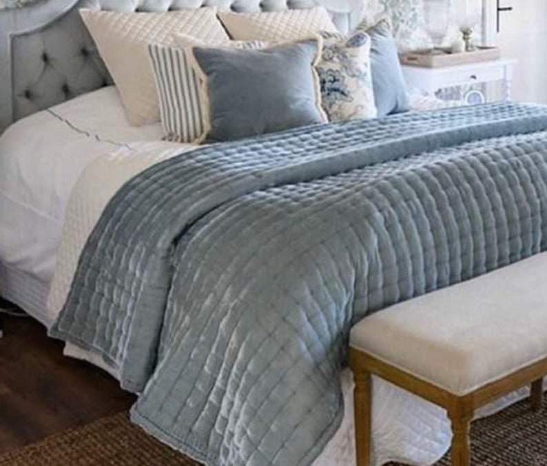 May include: A light blue quilted bedspread with a white bed and a white bench at the foot of the bed. The bed is made with white and blue patterned pillows.