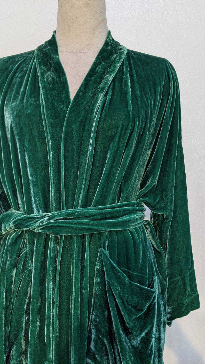 May include: A dark green velvet robe with a shawl collar and a tie belt. The robe has two large pockets on the front.