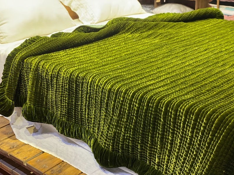 May include: A green velvet quilted throw blanket with a ruffled edge. The blanket is folded and displayed on a bed.