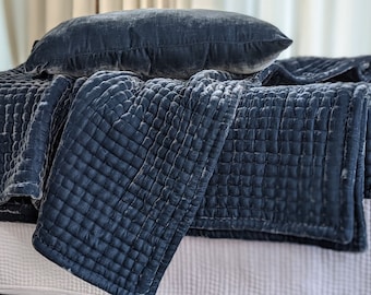 May include: A dark blue velvet quilted throw blanket with a matching pillow. The blanket is folded and draped over a white bed.