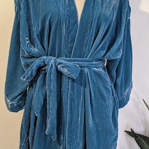 May include: A teal blue velvet robe with a self-tie sash. The robe has a V-neck and long sleeves.